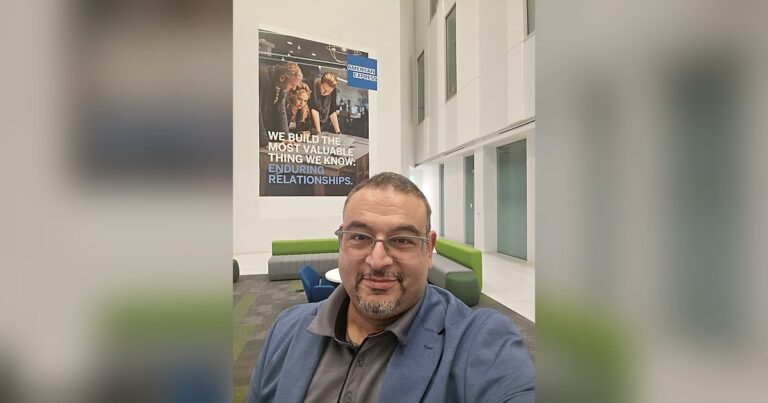 Sam Abraham at the American Express Sunrise campus in front of a poster reading 'We build the most valuable thing we know: Enduring Relationships.'